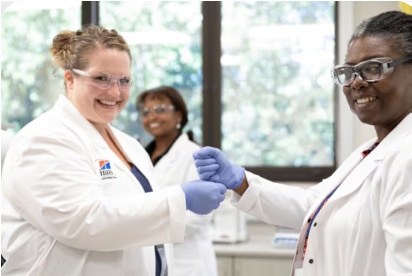 Drei lächelnde Frauen, Wissenschaftlerinnen in weißen Kitteln und Schutzbrillen